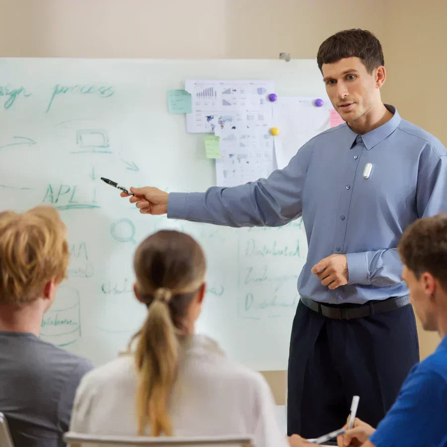 A man wearing a silver Plaud NotePin on his shirt while presenting at a whiteboard during a meeting.