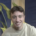 A smiling young man in a light-colored sweatshirt against a blue background with a plant.