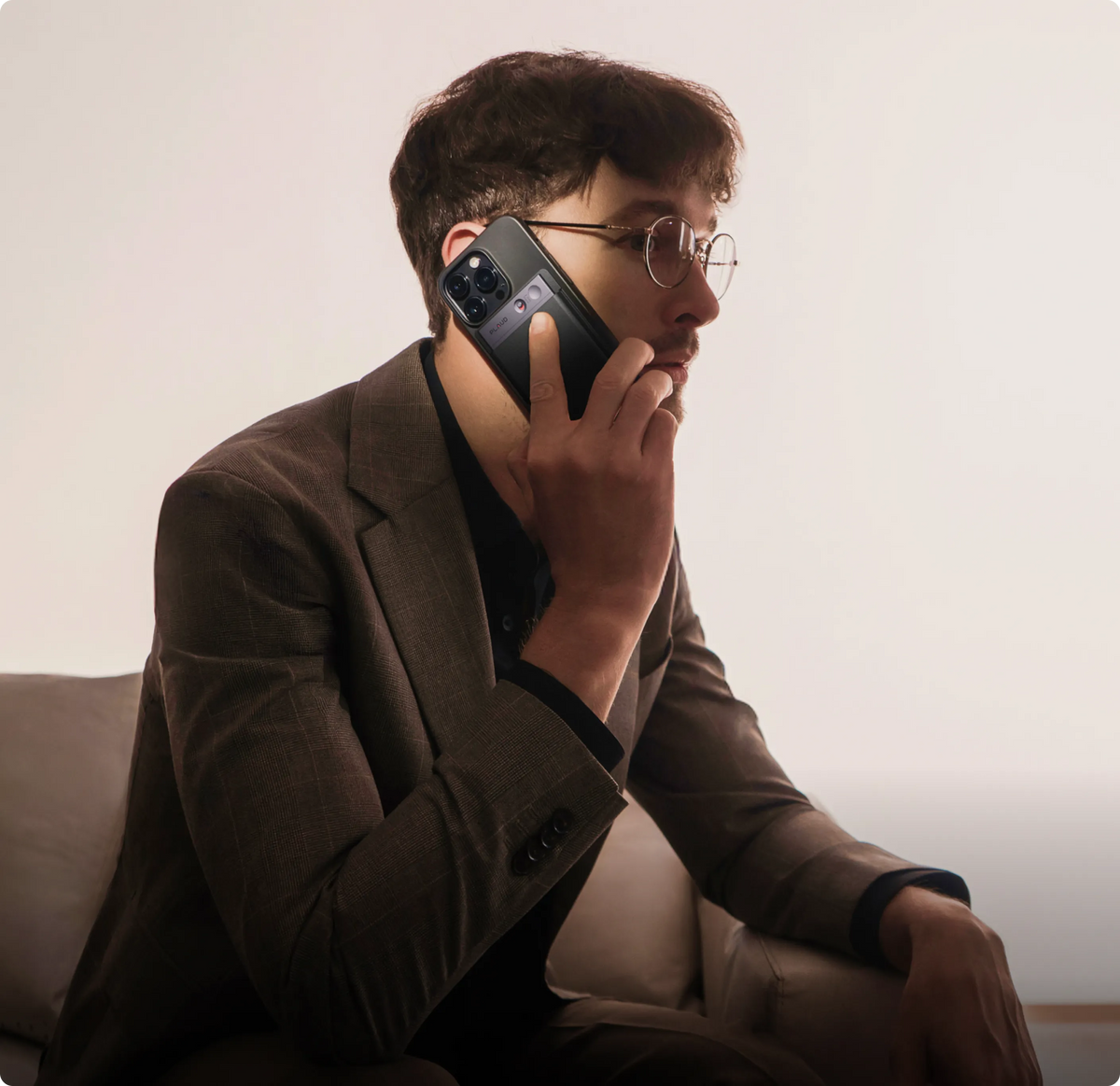 A man in a brown suit and glasses sits on a couch, holding a smartphone with a black Plaud Note AI voice recorder attached to the back.