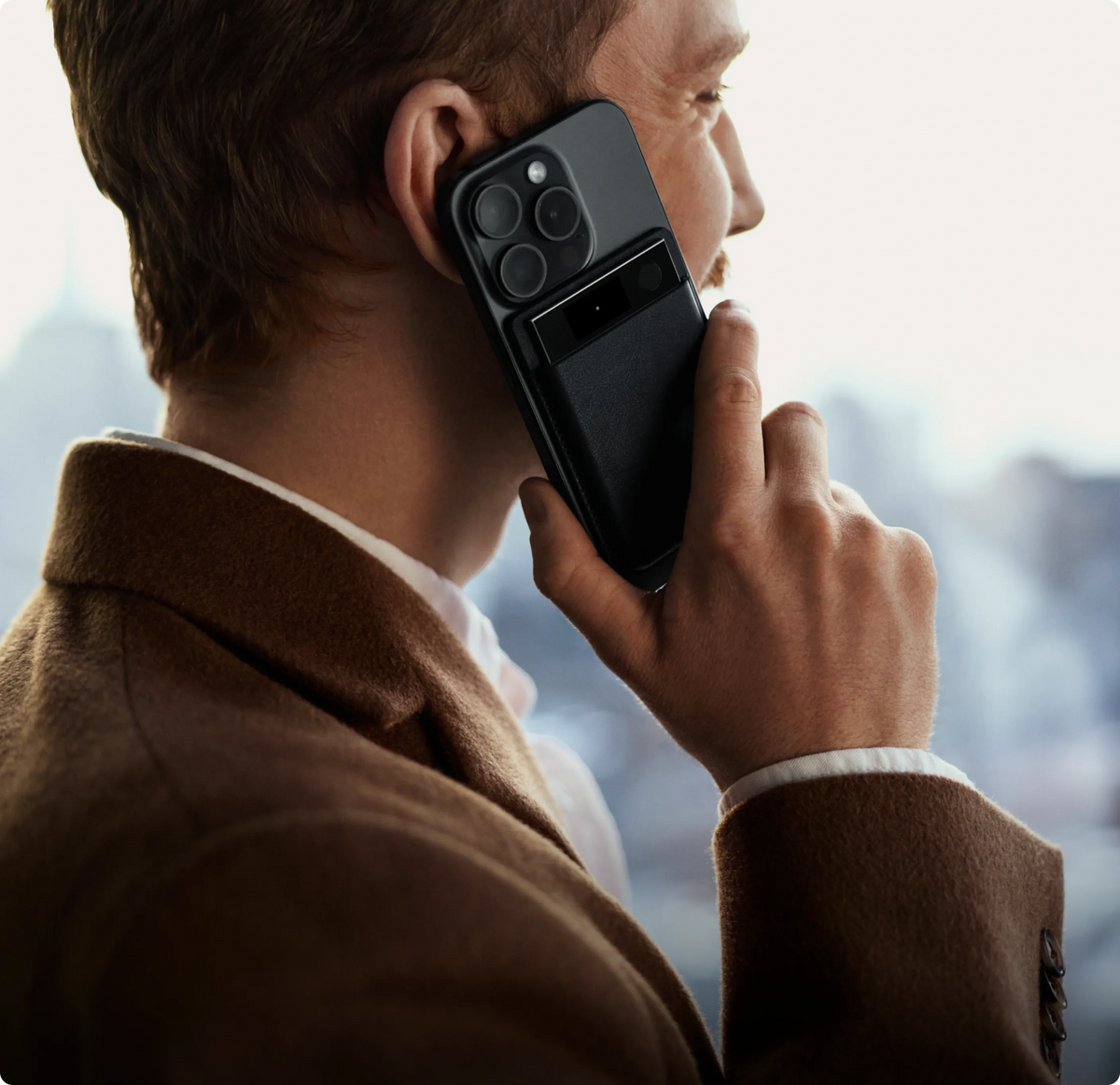 A man in a brown coat holds a smartphone to his ear with a black Plaud Note Pro AI voice recorder attached to the back.