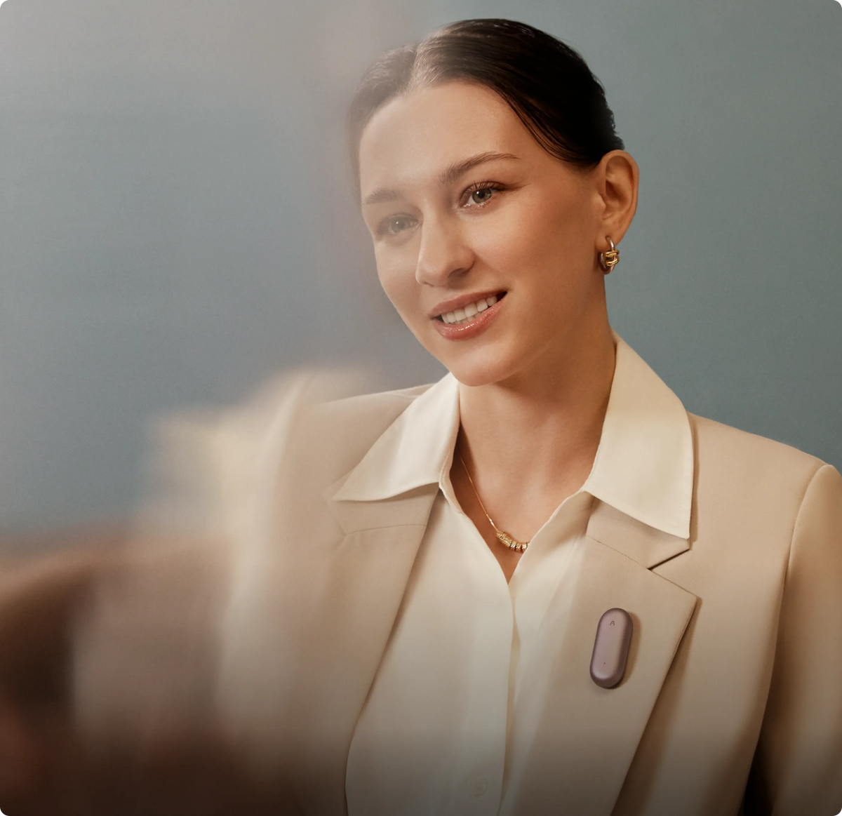 A smiling woman in a beige blazer and white shirt wears a lavender Plaud NotePin clipped to her lapel.
