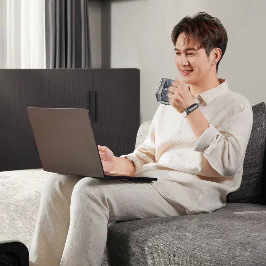Man on a sofa using a laptop and wearing a black Plaud NotePin on a white wristband.