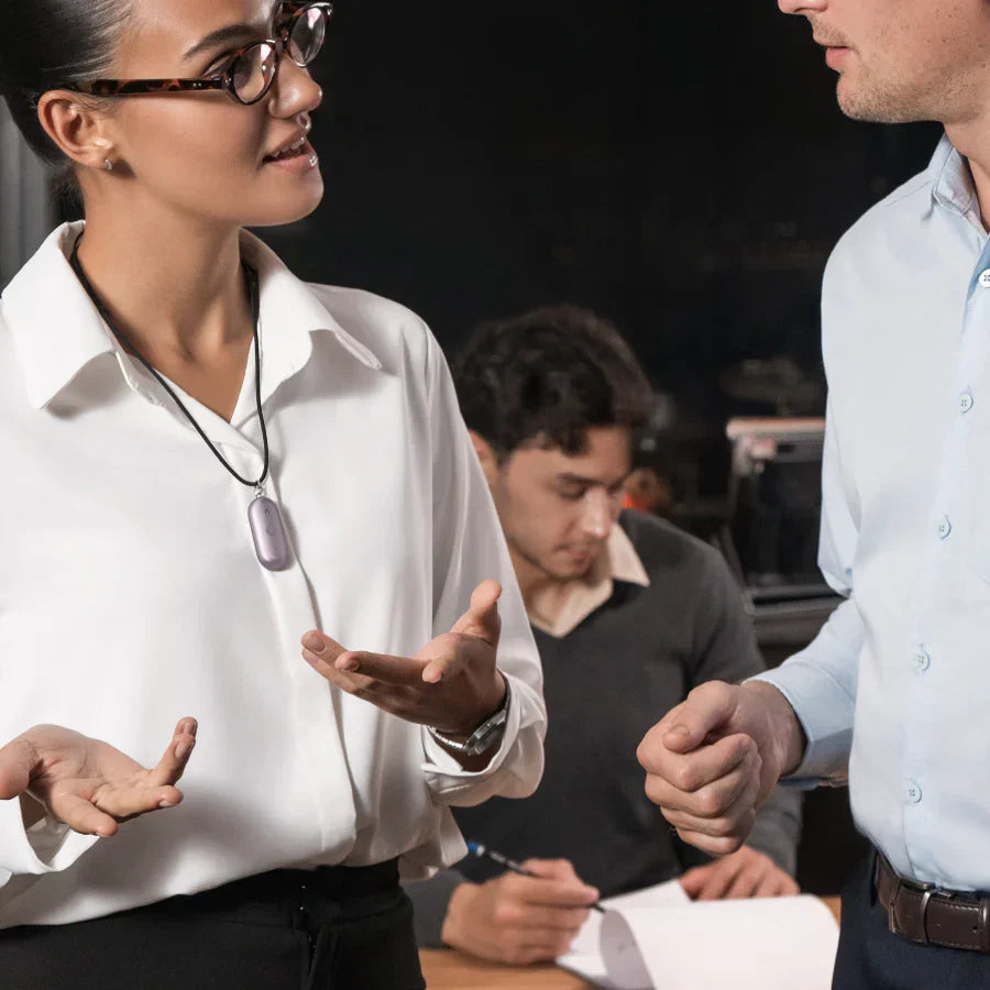 A woman wearing a purple Plaud NotePin on a lanyard while speaking with a colleague in a professional setting.