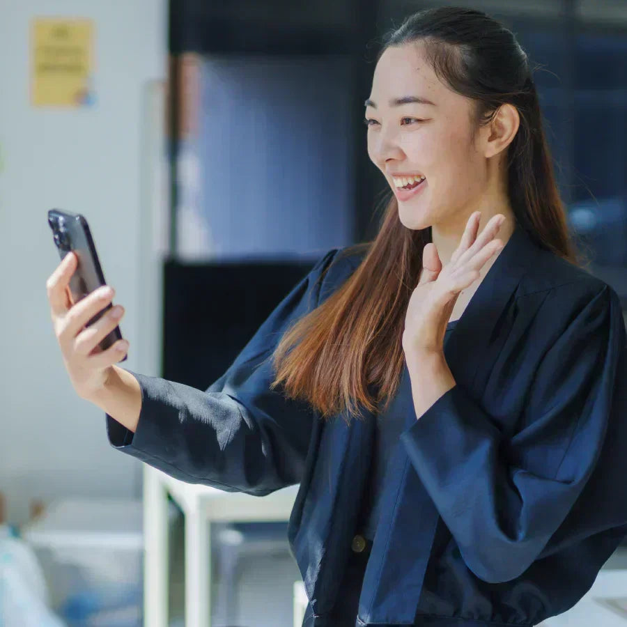 A woman waves during a video call on a phone with a black Plaud Note attached to the back.