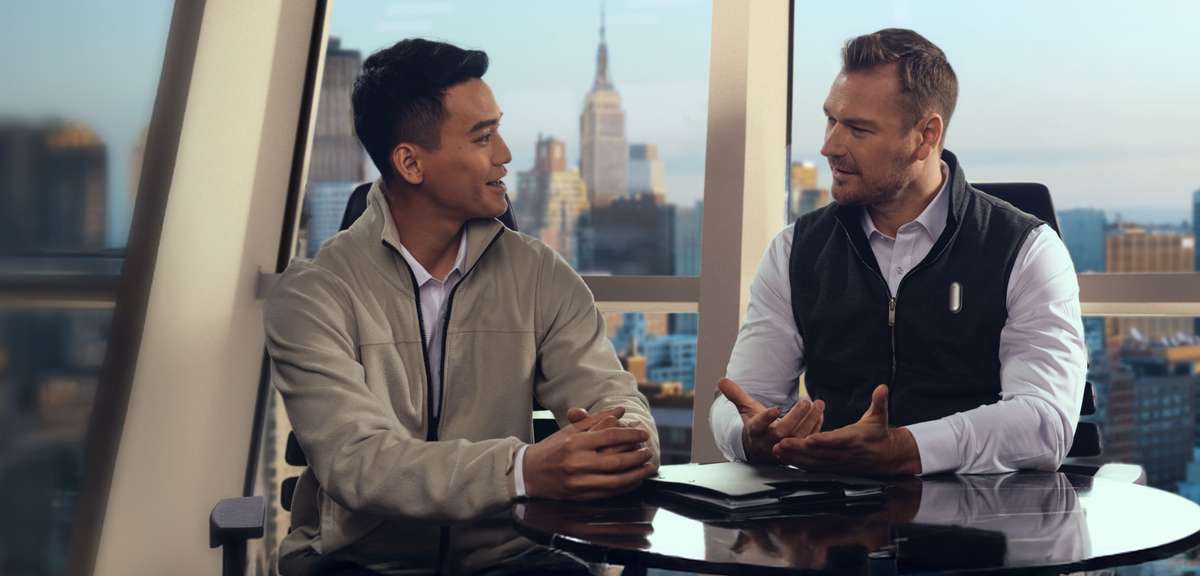 A man wearing a silver Plaud NotePin on his vest speaks with a colleague in a high-rise office overlooking a city.