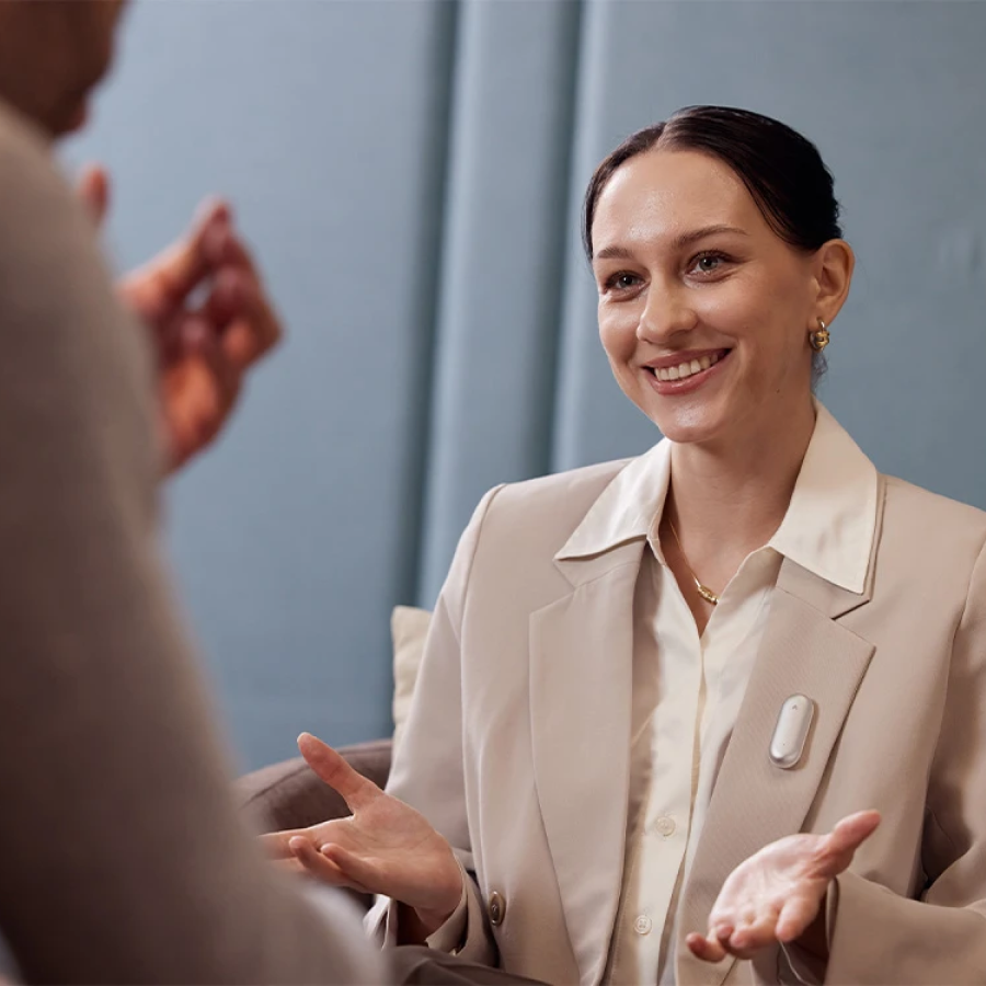 Smiling woman wearing a silver Plaud NotePin on her blazer lapel during a conversation.