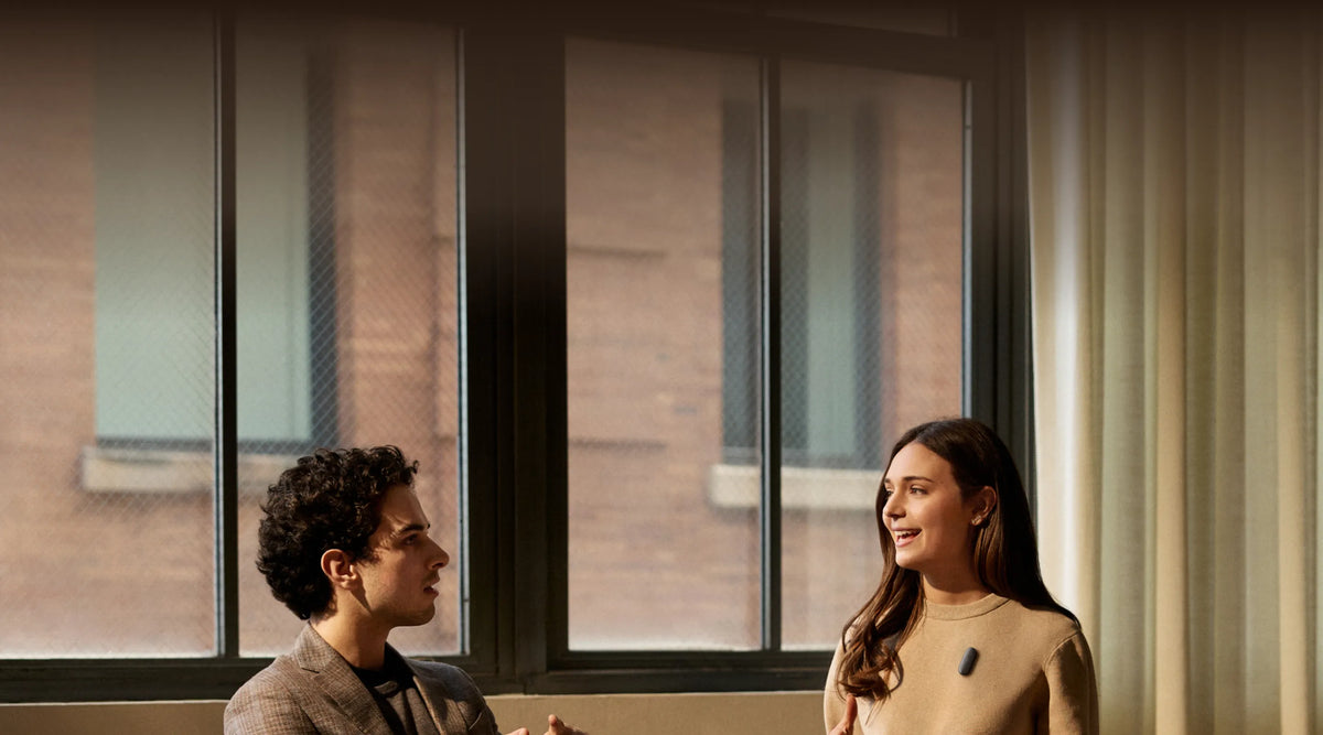 A man and a woman talk in front of a window while she wears a black Plaud wearable AI recorder on her beige sweater.