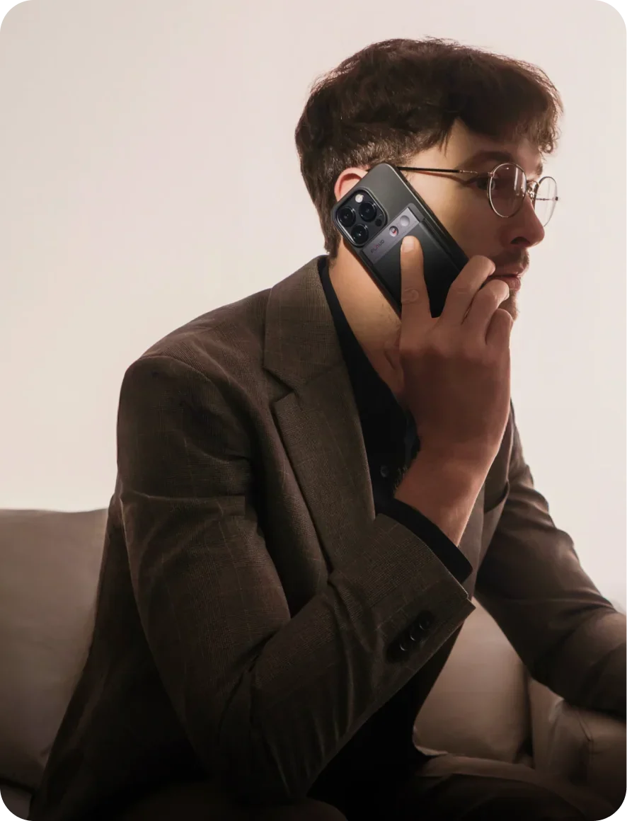 A man in a brown suit holds a smartphone with a black Plaud Note attached to the back, his finger resting near the recording button.
