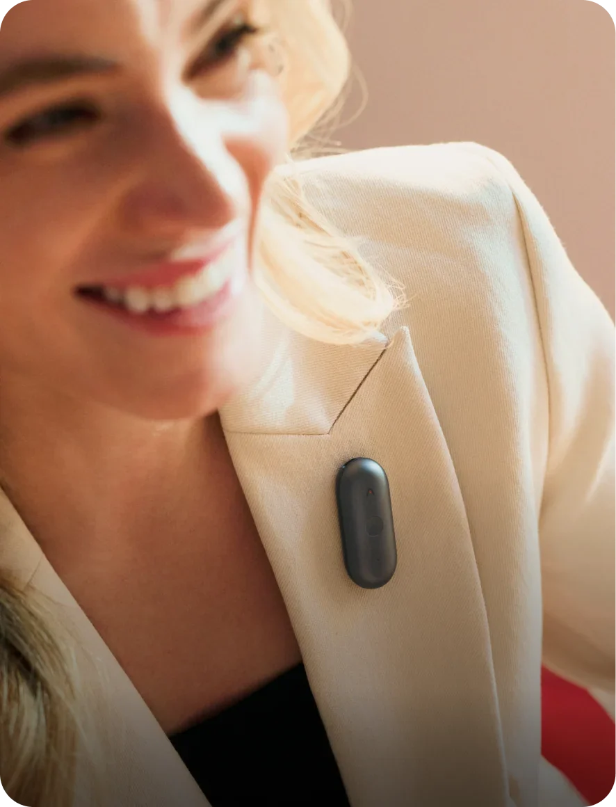 A smiling woman wears a dark grey Plaud NotePin S clipped to the lapel of her cream blazer, showcasing the wearable AI recorder.
