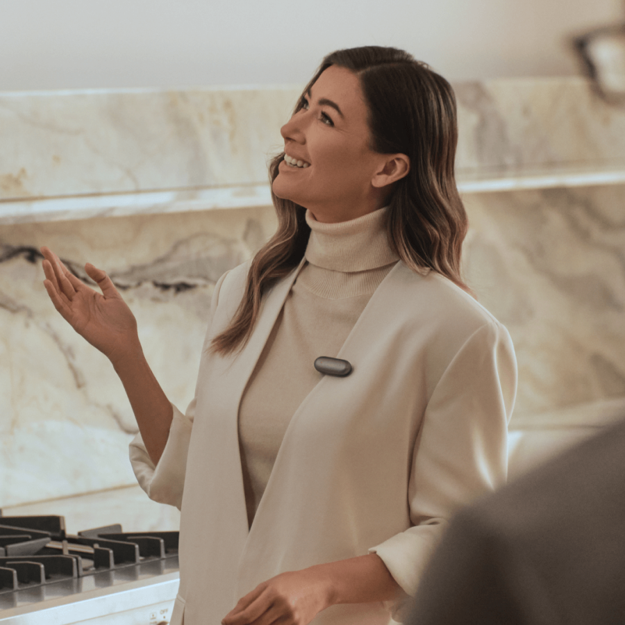 A woman wearing a Cosmic Gray Plaud NotePin on her white blazer while gesturing during a conversation.