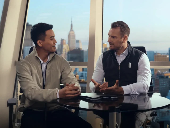Two men meeting in an office with a city view. One wears a silver Plaud NotePin on his vest.