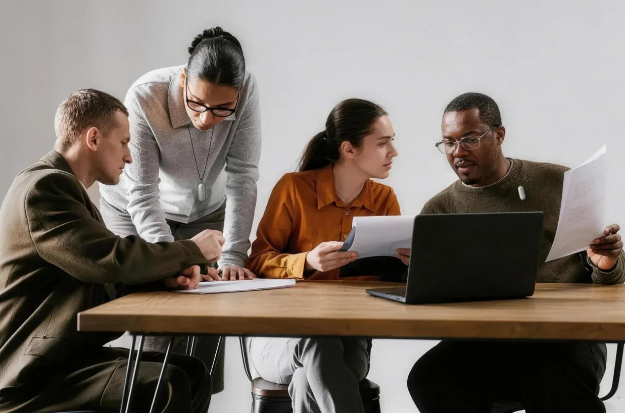 Four colleagues discuss documents around a laptop at a wooden table, with two members wearing silver Plaud wearable AI recorders.