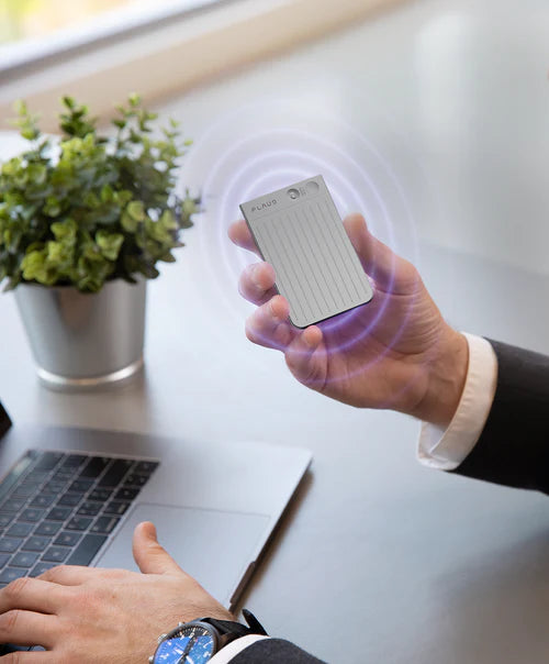 A person holds a silver Plaud Note Pro with purple soundwave graphics at a desk with a laptop.