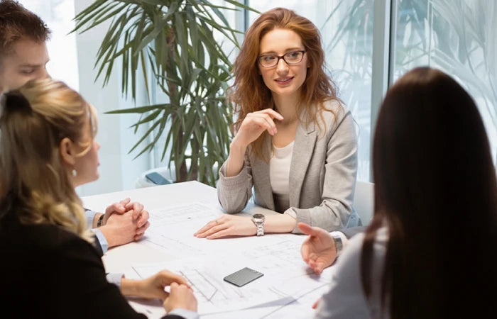 A black Plaud Note sits on a conference table during a business meeting with four professionals discussing documents.