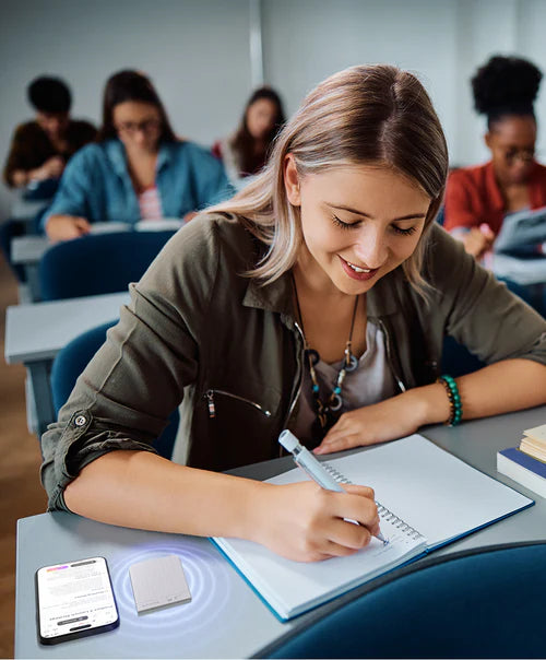 A student uses a silver Plaud Note Pro to record a lecture while taking handwritten notes in a classroom.